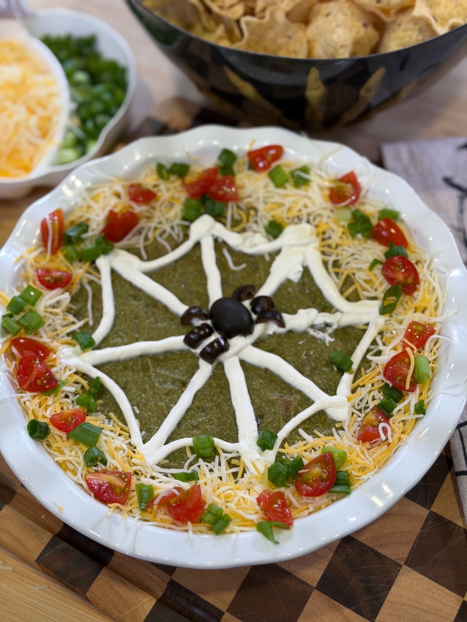 A Halloween-themed taco dip in a white dish, decorated with a sour cream spiderweb and an olive spider, surrounded by shredded cheese, chopped green onions, and cherry tomatoes.